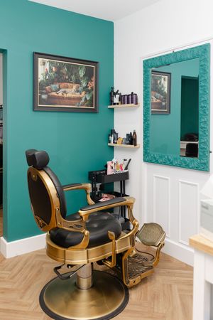 Elegant gold and black barber chair in a teal-walled salon with herringbone floors, ornate mirror, and product shelves.