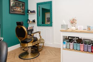 Elegant barber shop interior with gold vintage chair, teal walls, herringbone floor, and colorful product display shelves.