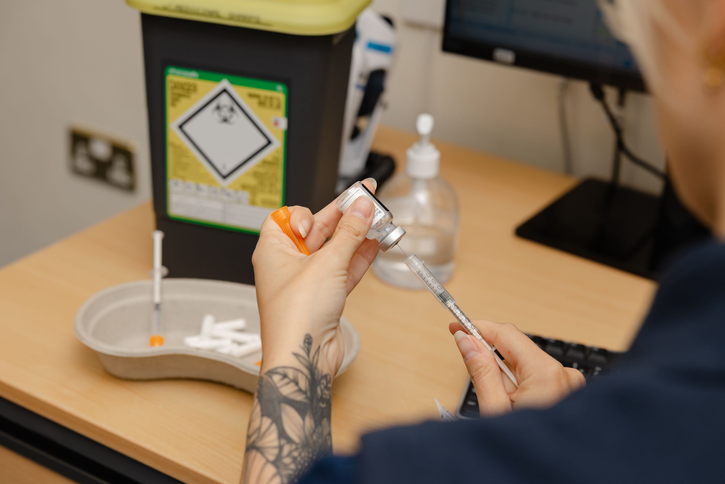 Healthcare worker with tattoos preparing a vaccine syringe at a medical workstation with sharps disposal bin.
