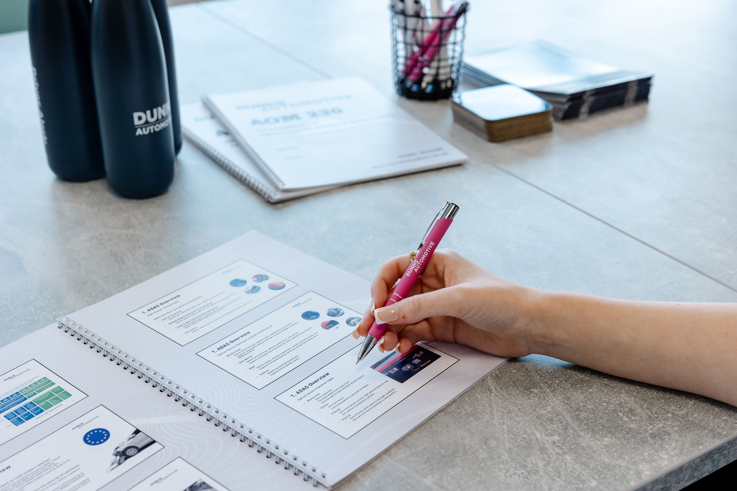 A person writing in a spiral notebook with a pink pen on a desk with documents and a water bottle.