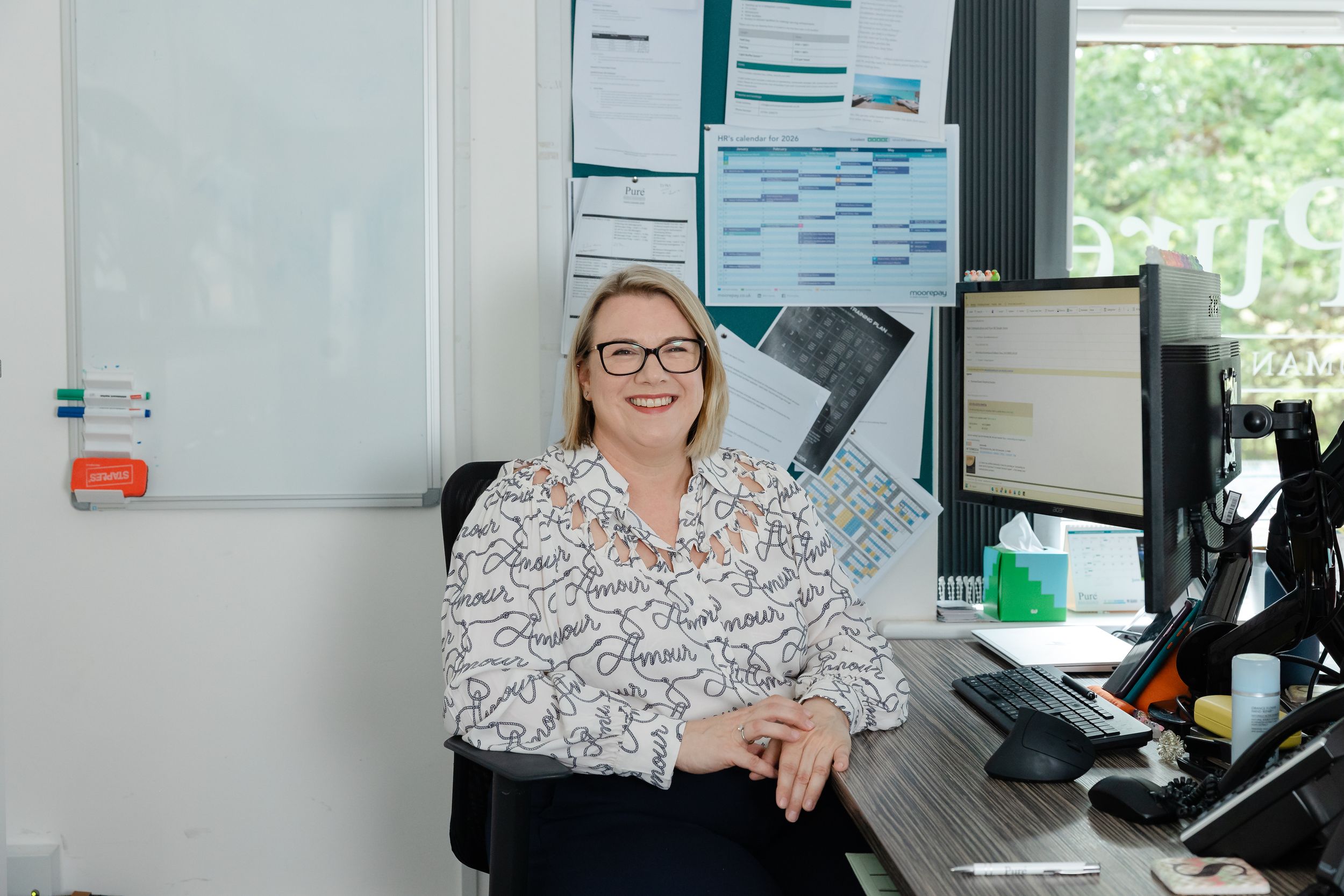 Woman in patterned blouse smiling at desk in modern office with whiteboard and computer monitors.