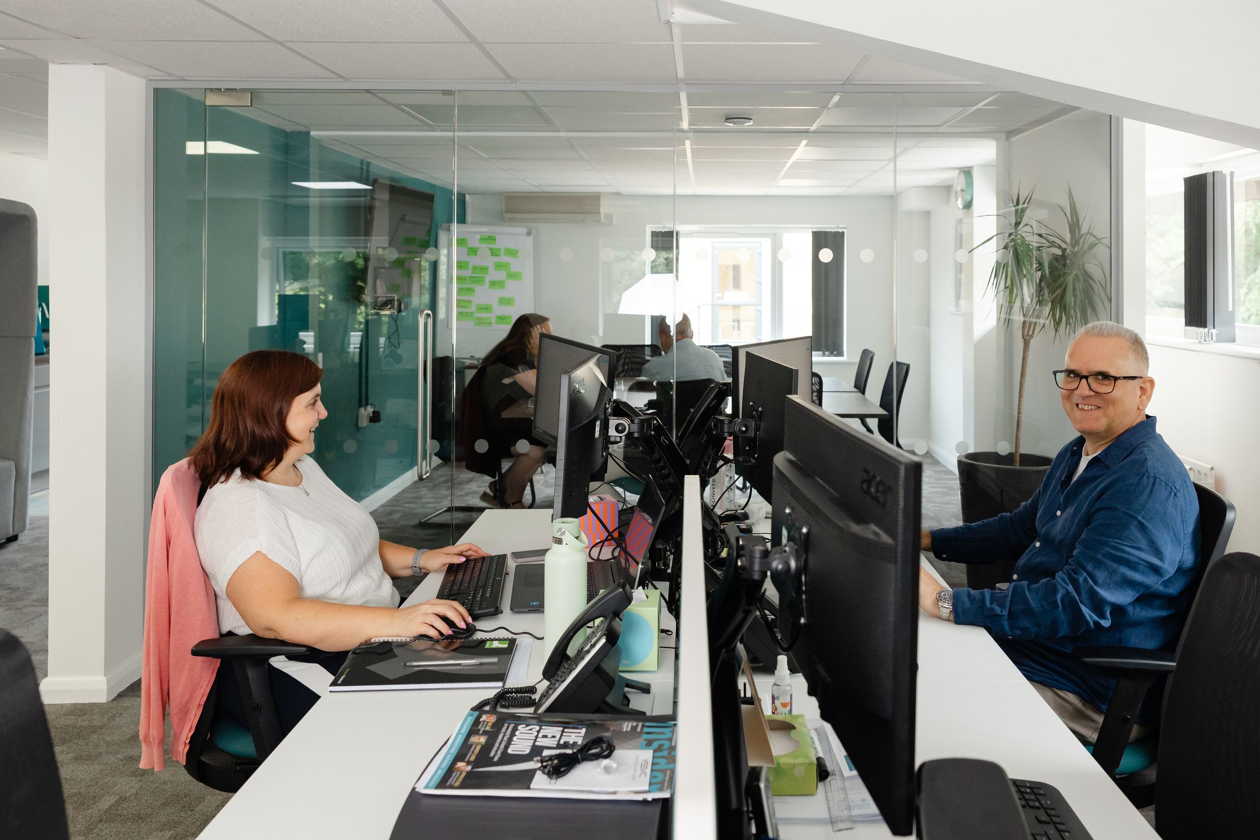 Two office workers smile at their desks with multiple monitors in a modern open-plan workspace with glass partitions.