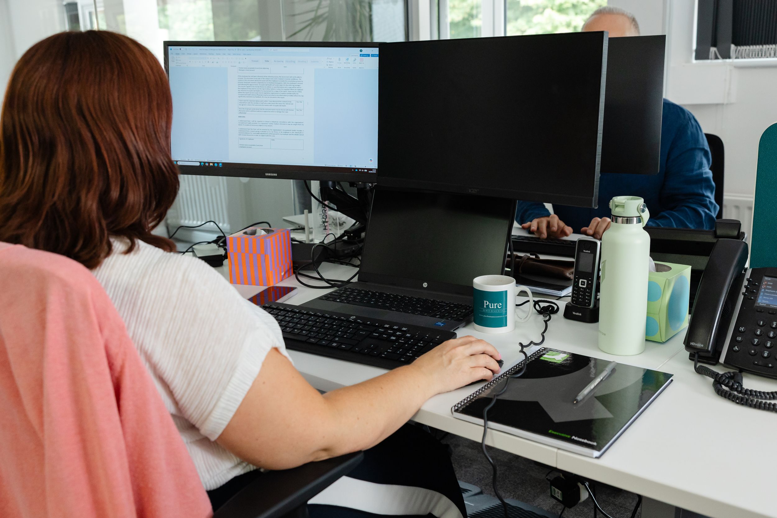 Woman working at dual monitor desktop computer setup in modern office workspace with colleague in background.