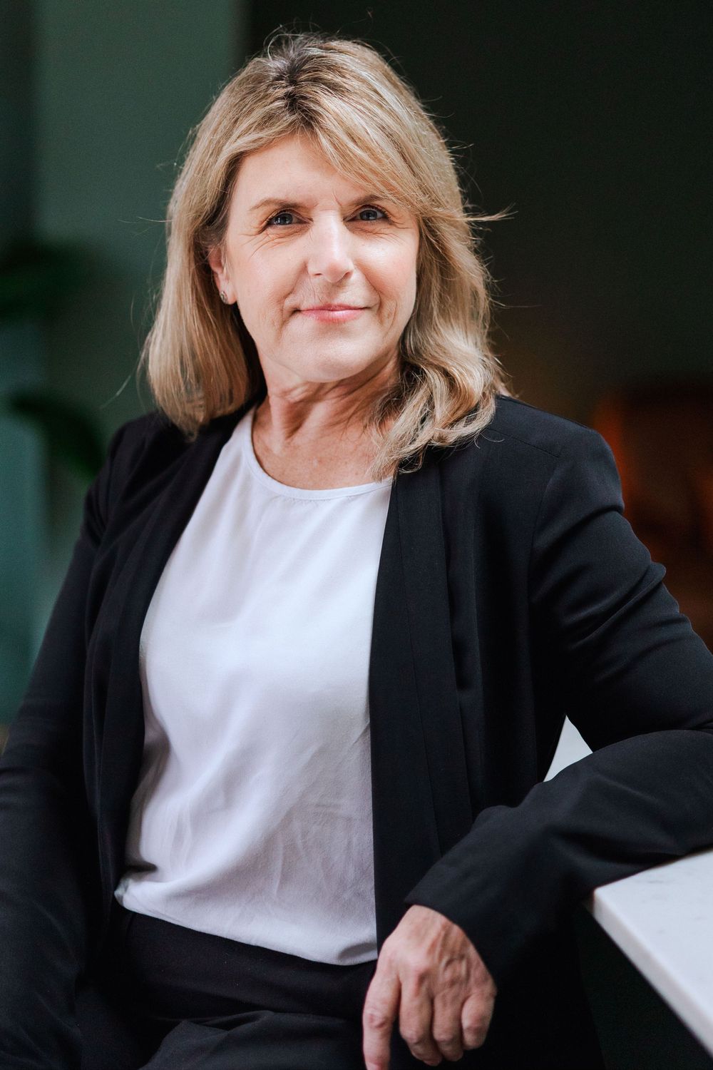 Professional portrait of someone wearing a black blazer and white top while sitting casually in a business setting.