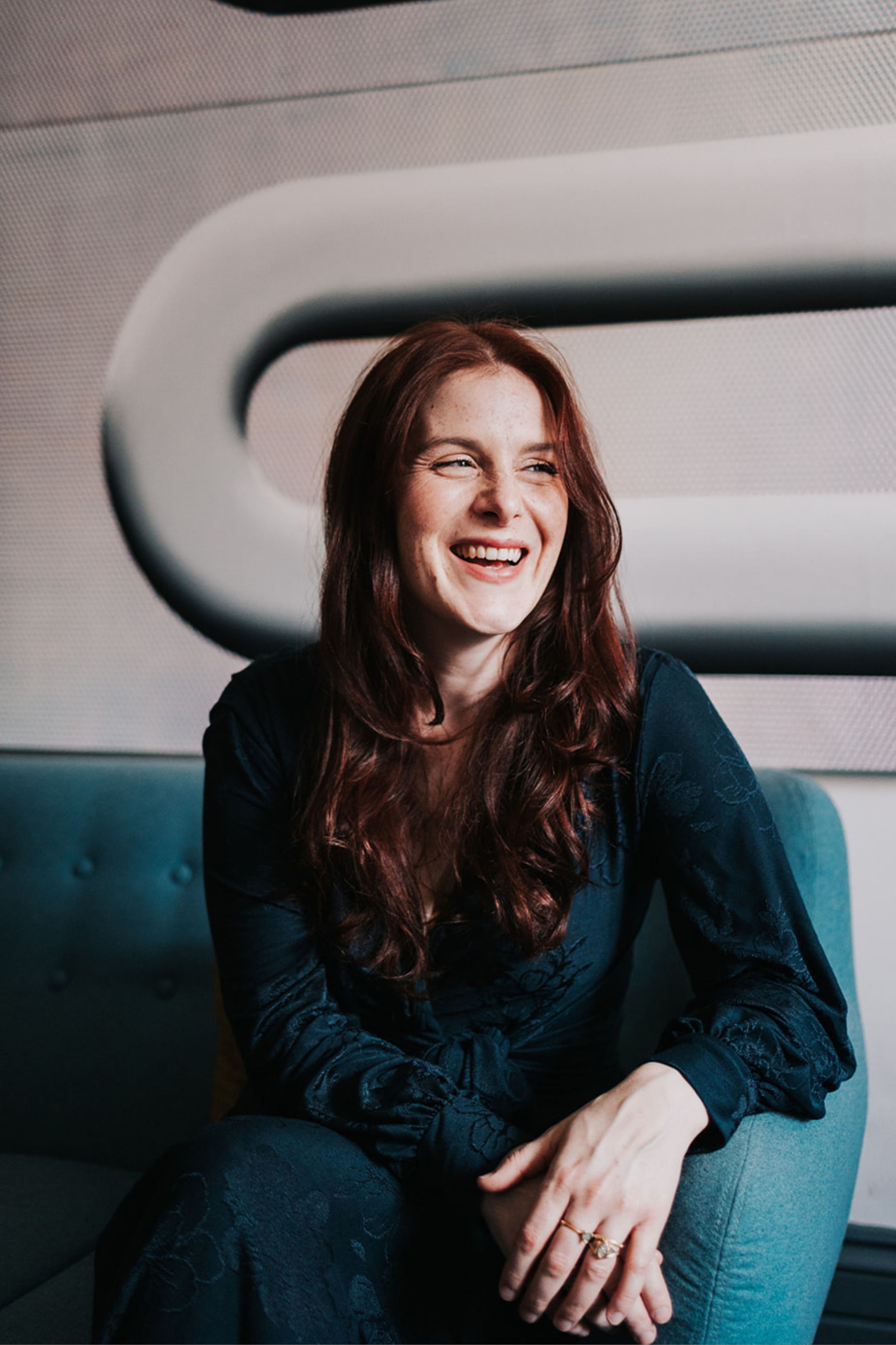 A joyful candid portrait shows someone with long red hair laughing while seated on a blue couch.
