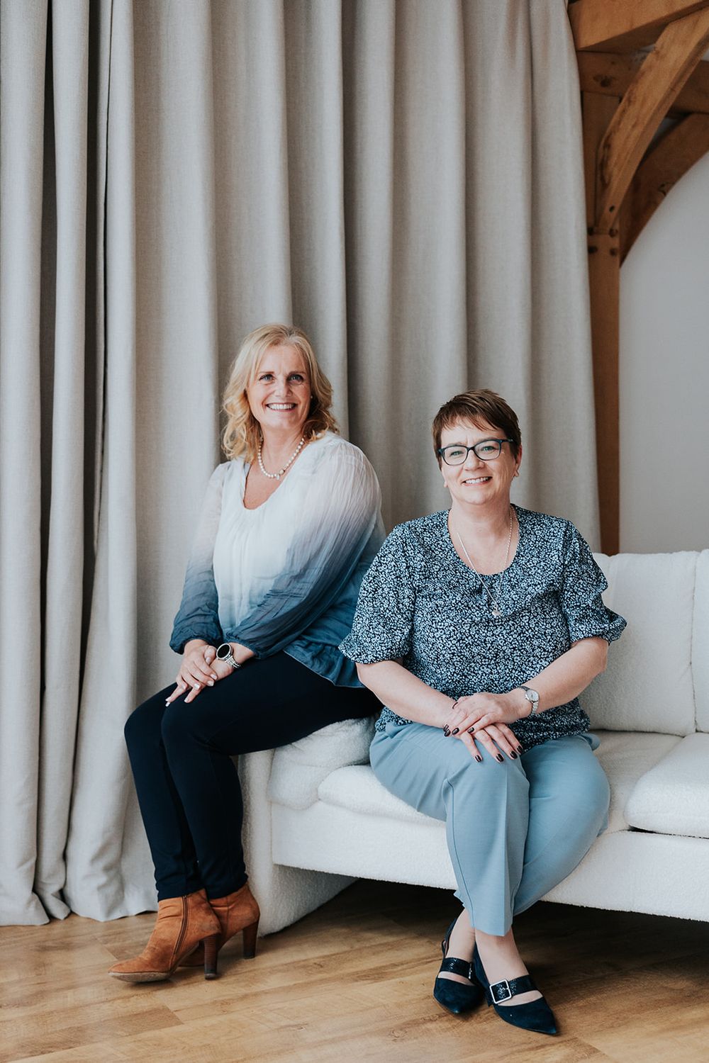 Two women sit comfortably on a white couch against light curtains in a room with wooden flooring and accents.