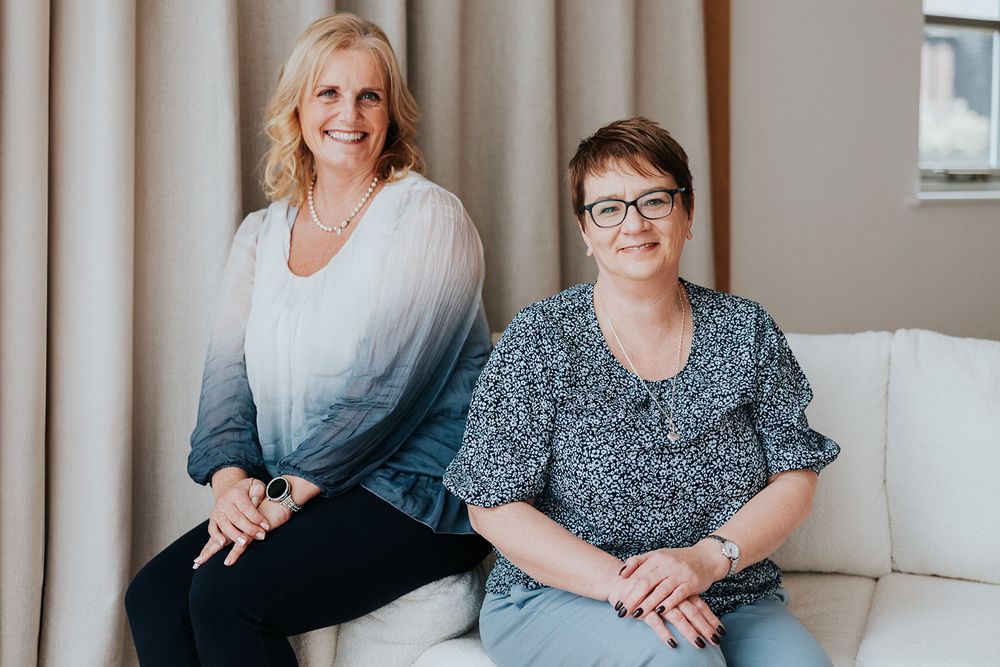 Two people sitting together on a beige couch, one wearing white ombre top and one in a floral print blouse.