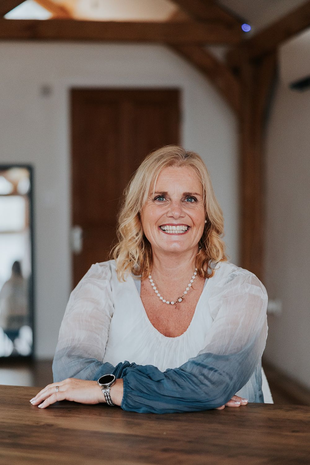 A person in a white and blue ombre blouse sits smiling at a wooden table while wearing a pearl necklace and watch.