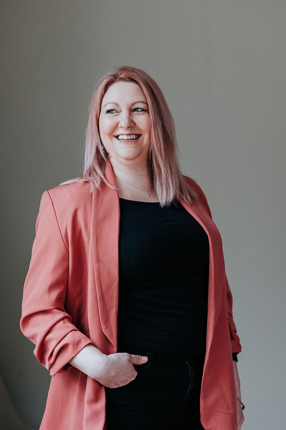 Professional portrait of a person in a coral blazer and black dress standing against a gray background smiling warmly.