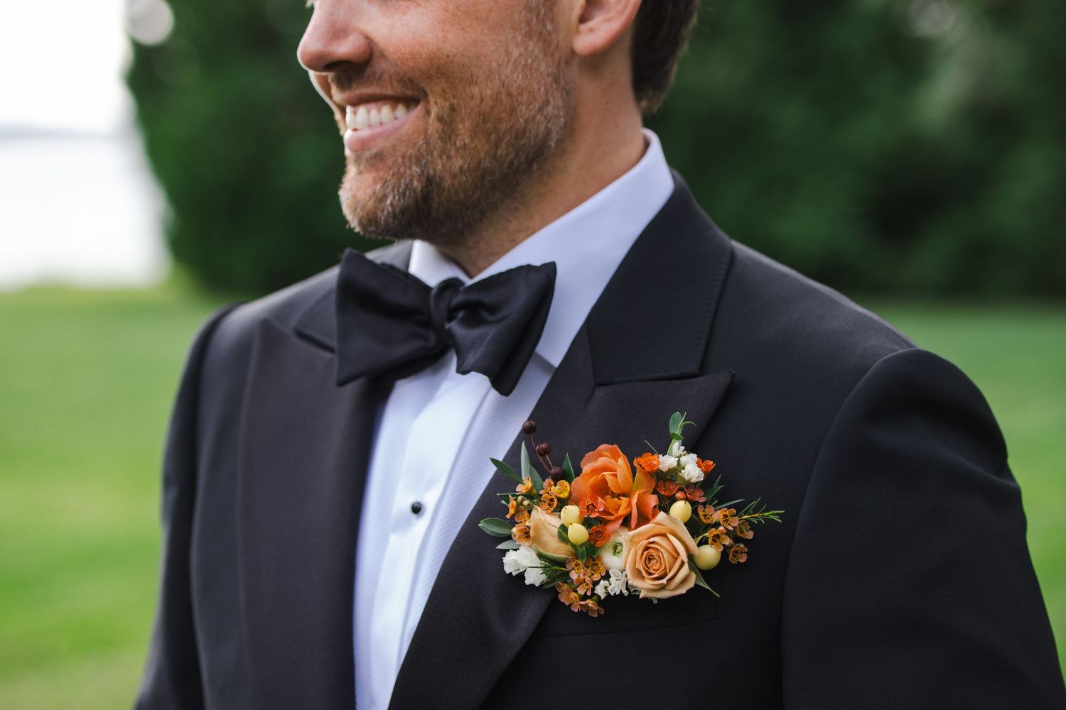 Close up of an orange and white floral boutonniere worn on a black tuxedo with bowtie.