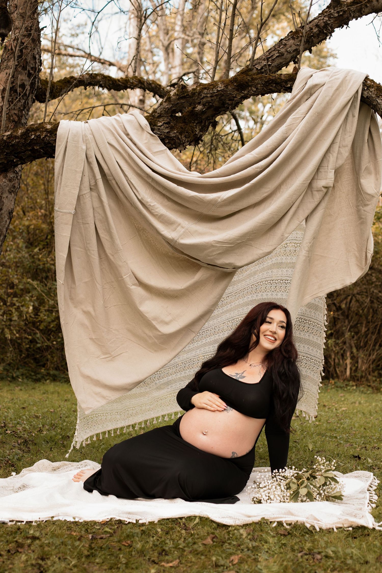 A maternity photo in autumn features a draped beige backdrop hanging from a tree branch with someone posing on a blanket below.