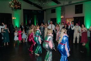 Party guests dance under green lighting and chandeliers at a wedding reception.