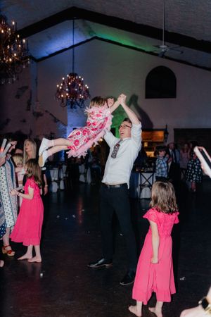 A joyful dance scene at a wedding reception features playful twirls under ornate chandeliers in a dimly lit venue.