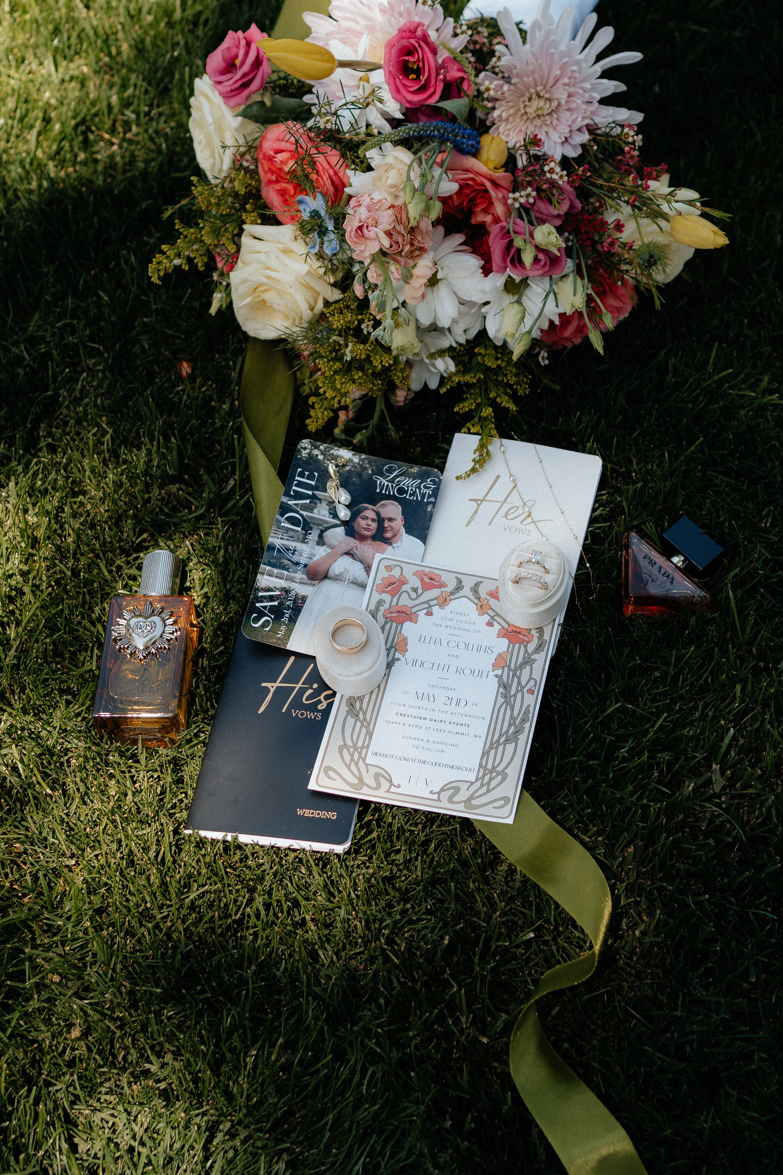 Wedding flat lay with colorful bridal bouquet, rings, perfume, invitations, and His & Hers books on green grass.