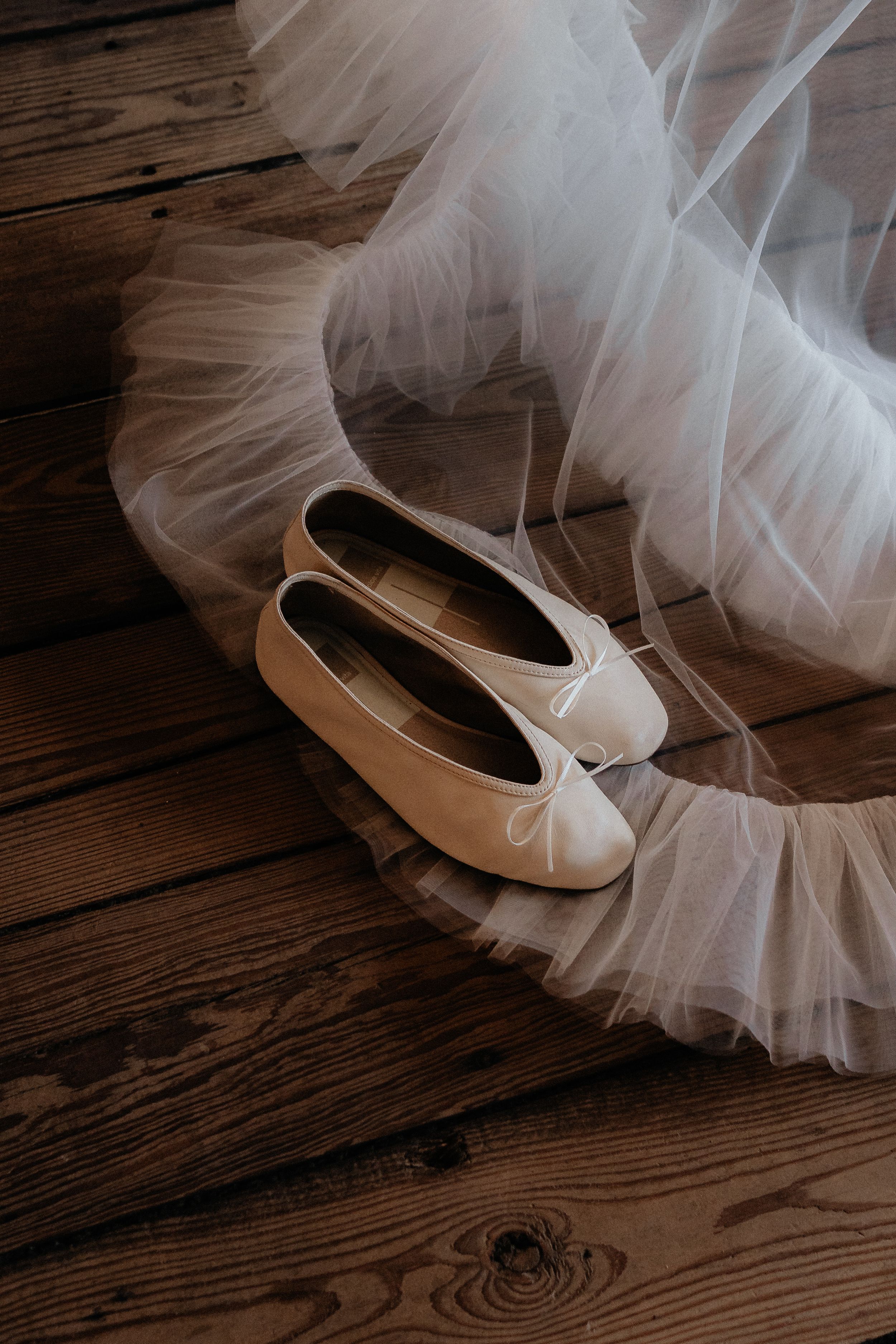 Ivory bridal ballet flats with bow detail resting on rustic wood floor beside flowing white tulle wedding dress.