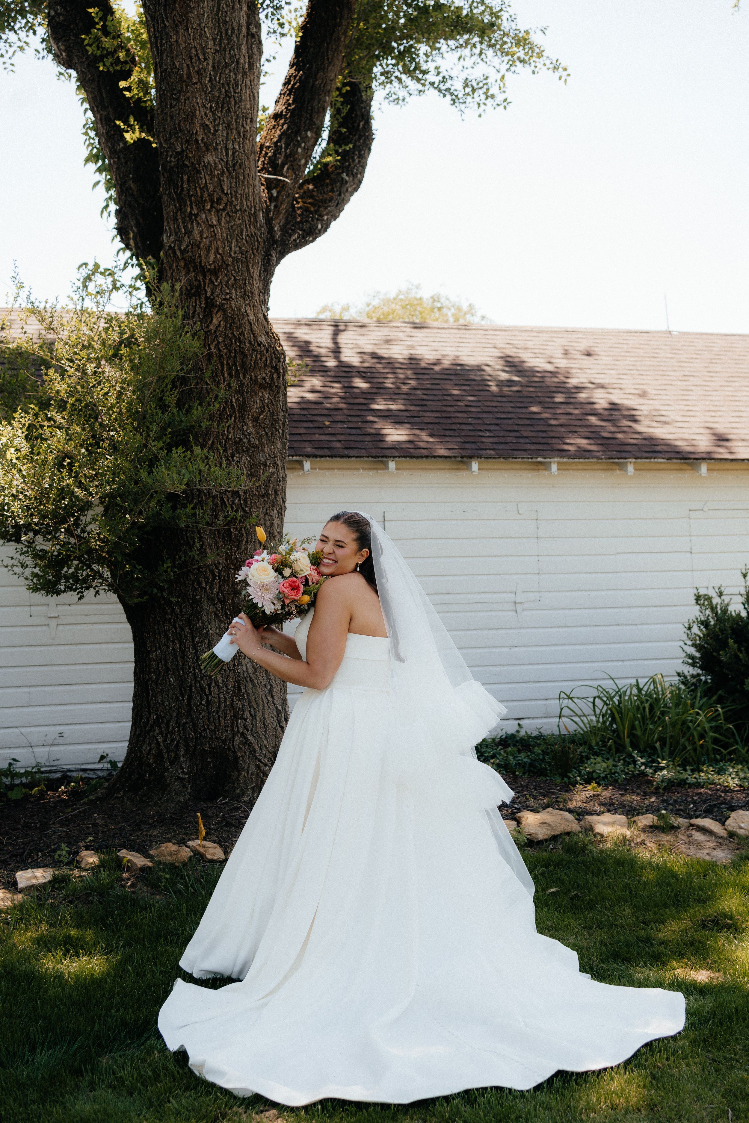 Bride in white gown and veil holds colorful bouquet, posing by large tree near white building on sunny day.