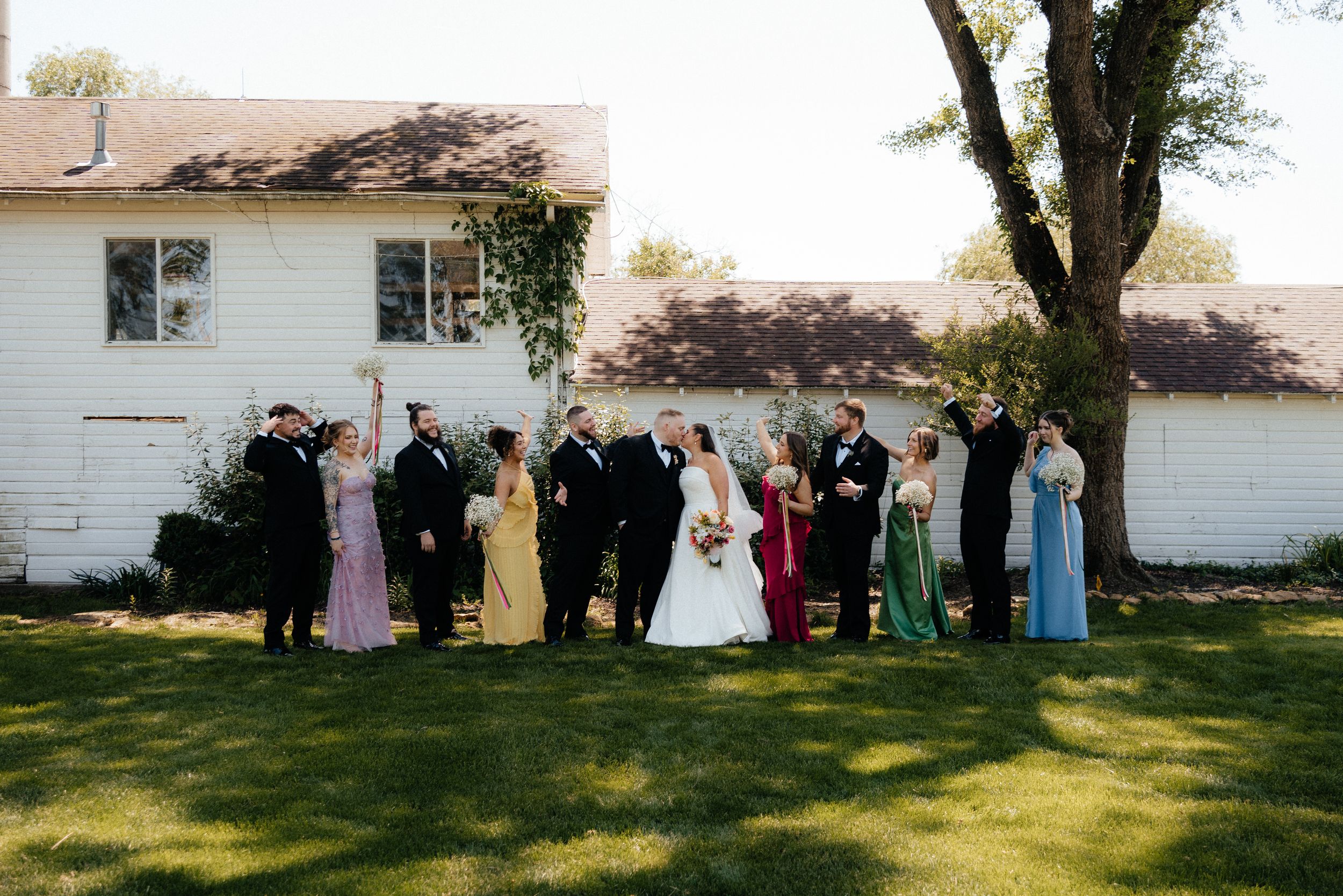 Wedding party in formal attire poses on green lawn beside white house, bridesmaids in colorful gowns and groomsmen in black tuxedos.