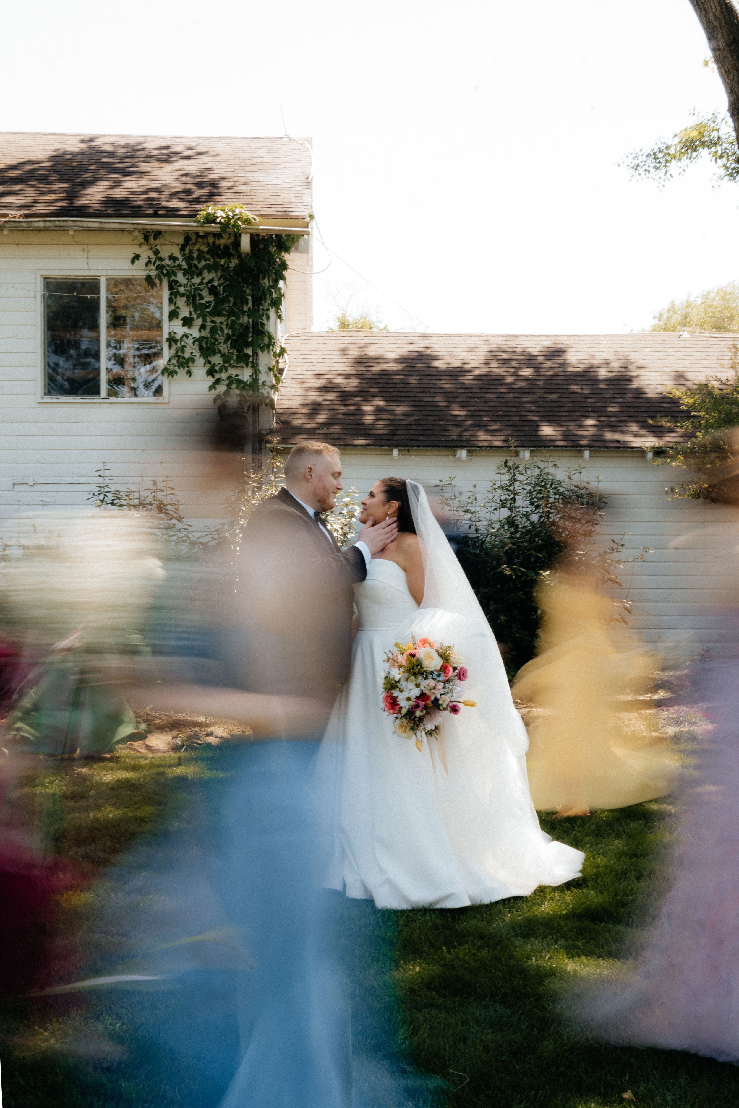 Bride and groom share a romantic moment surrounded by blurred wedding guests in a sunlit backyard setting.