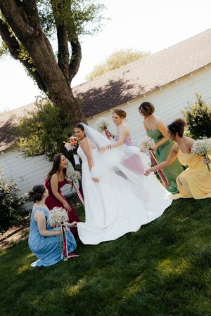 Bridesmaids in colorful dresses help arrange bride's white wedding gown train outdoors near white barn.