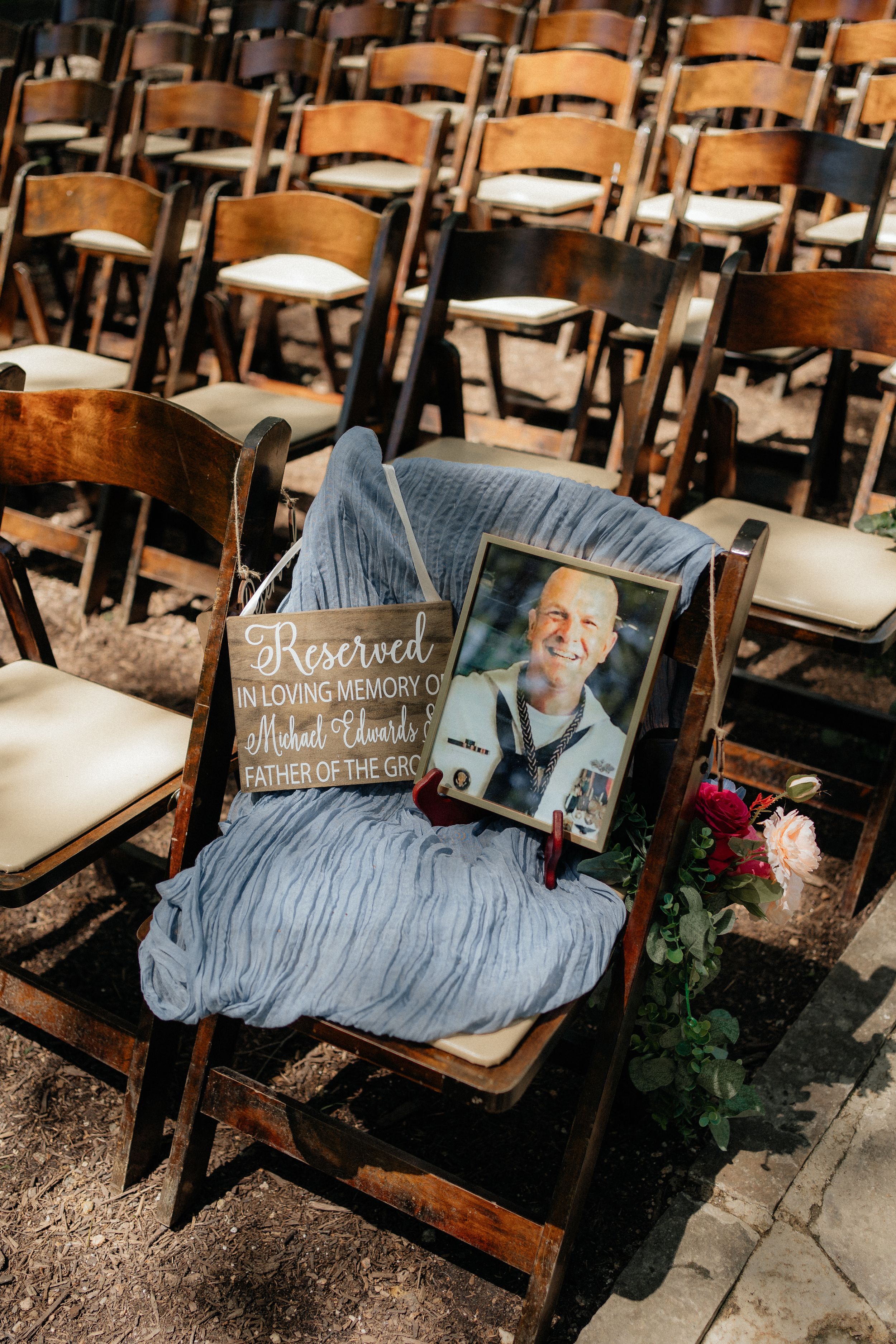 Memorial chair draped in blue fabric with a wooden sign and photograph at an outdoor wedding ceremony setup.