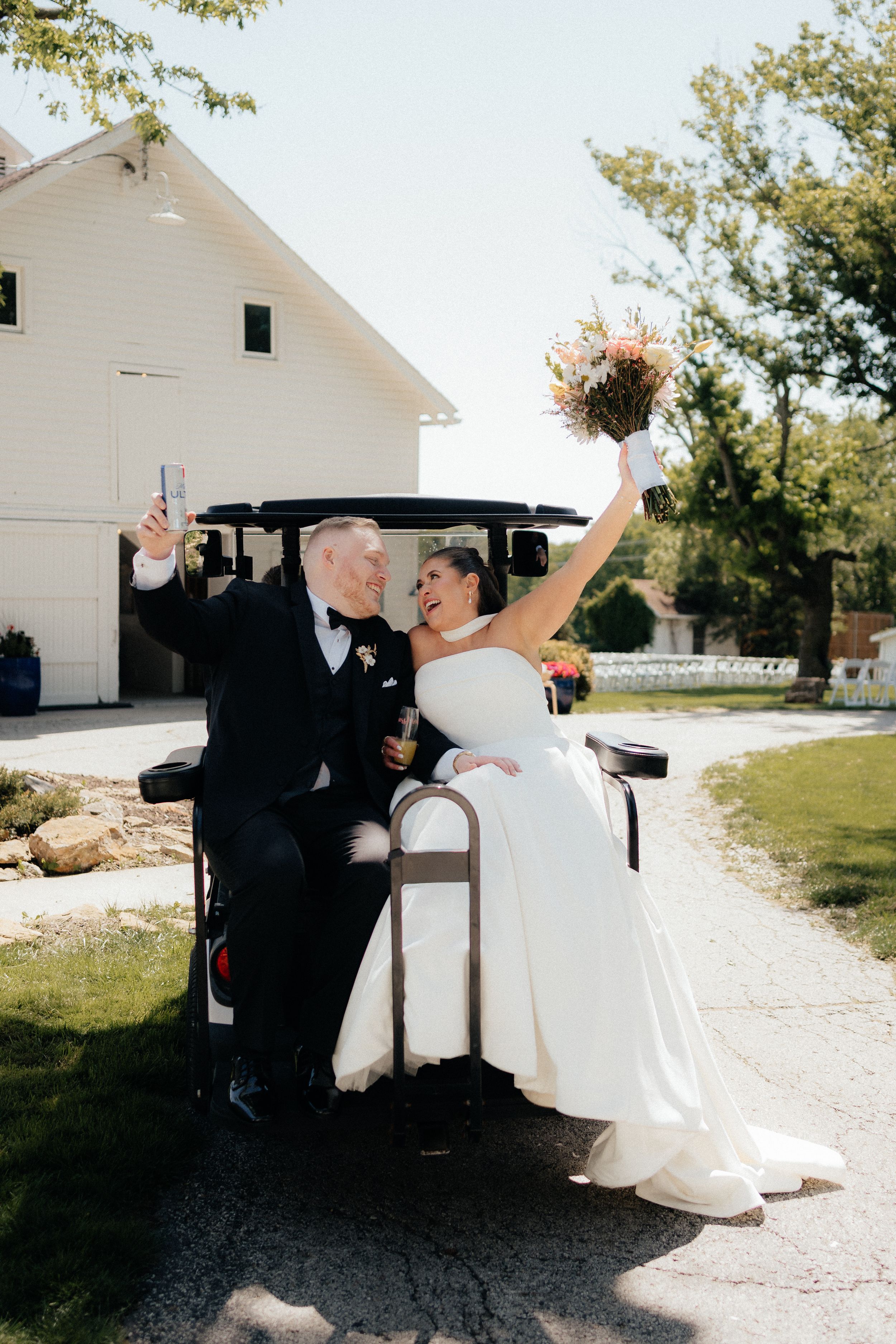 Joyful bride and groom celebrating on a golf cart outside a white barn, bride holding bouquet aloft on sunny wedding day.