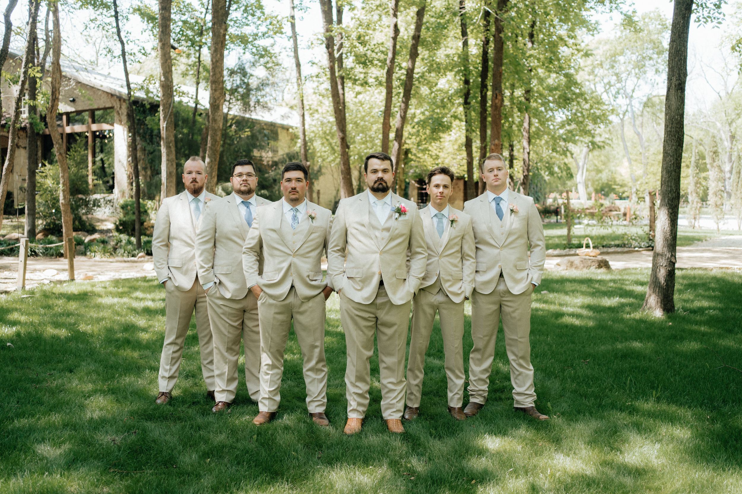 Groomsmen in tan suits standing together on green lawn in front of trees.