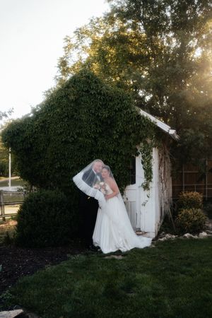 Bride and groom share a romantic kiss outdoors near an ivy-covered building at golden hour on their wedding day.