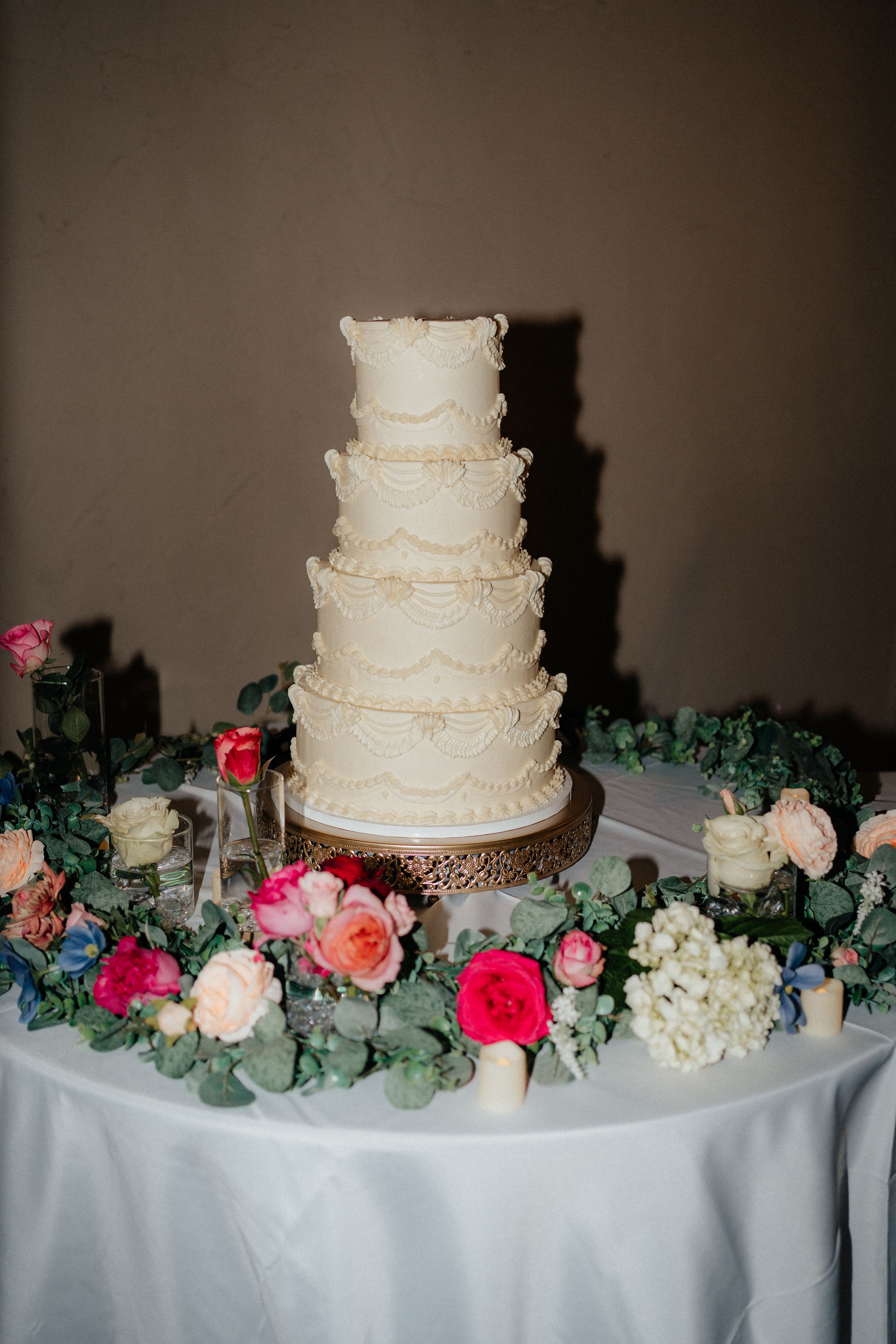 Elegant four-tier white wedding cake decorated with lace patterns and surrounded by colorful flowers.