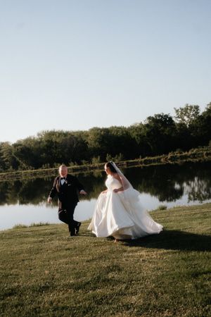 Bride in white gown and groom in black tux playfully run along a green lakeside lawn at golden hour.