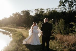 Bride and groom walking hand in hand along a sunlit waterway surrounded by lush greenery on their wedding day.