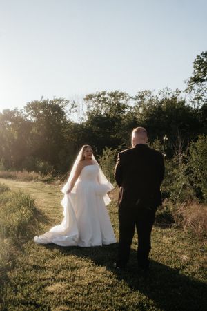 Bride in white gown and veil faces groom in black suit during golden hour outdoor wedding portrait.