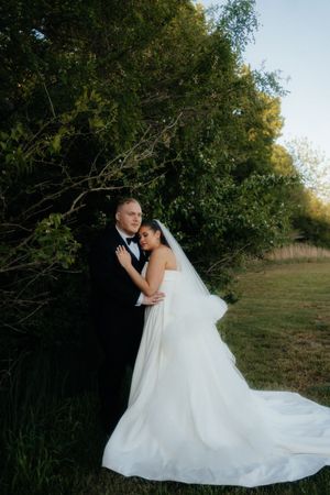 A bride in a white gown and veil embraces a groom in a black tuxedo outdoors near lush green trees.