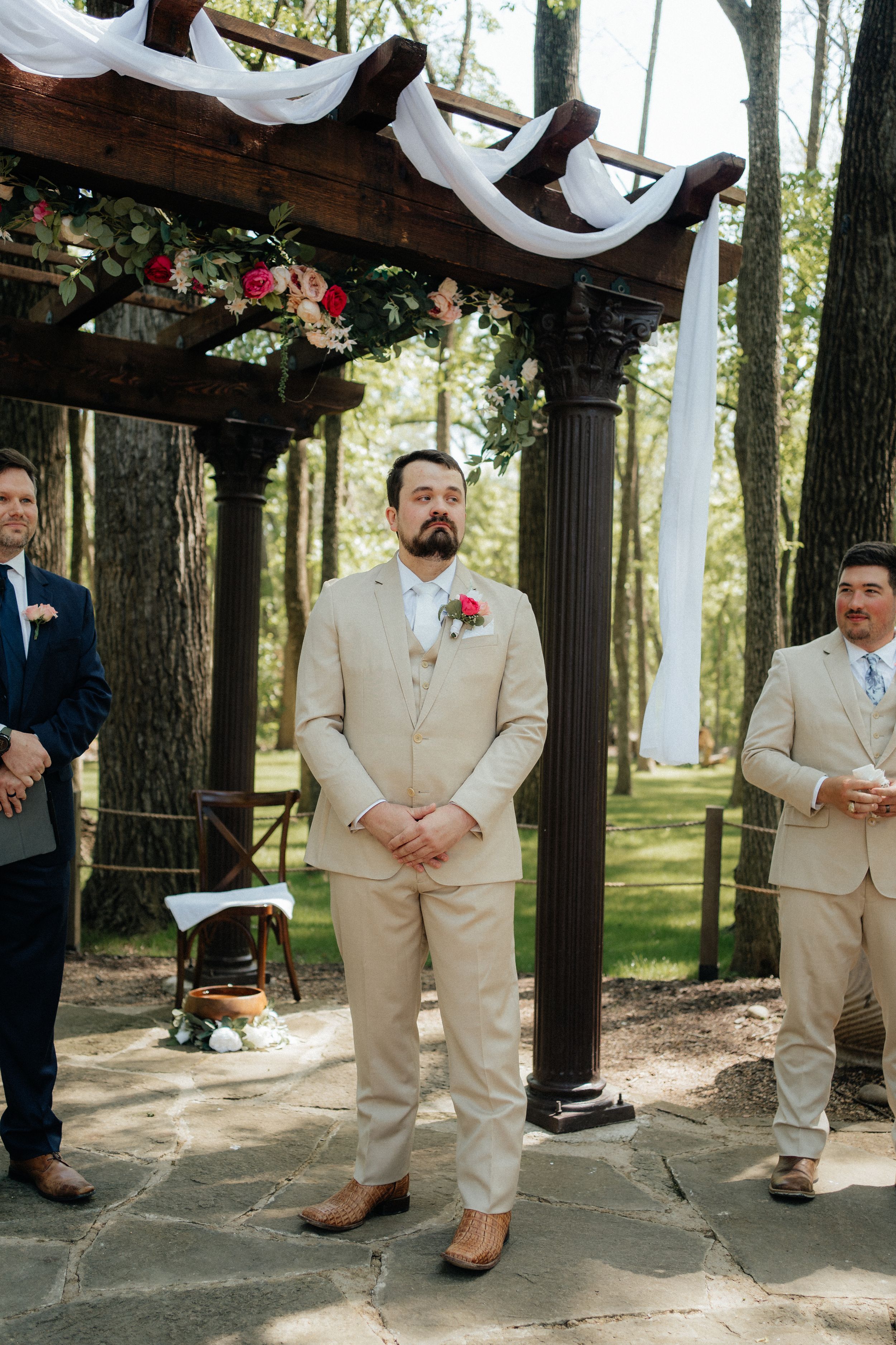 Wedding ceremony setup with decorated wooden arch and groomsmen standing under draped fabric.