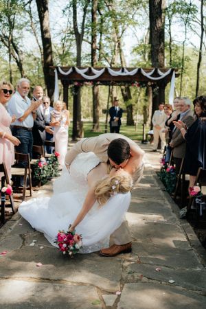 Romantic dip kiss between newlyweds under decorated arch at outdoor woodland ceremony.