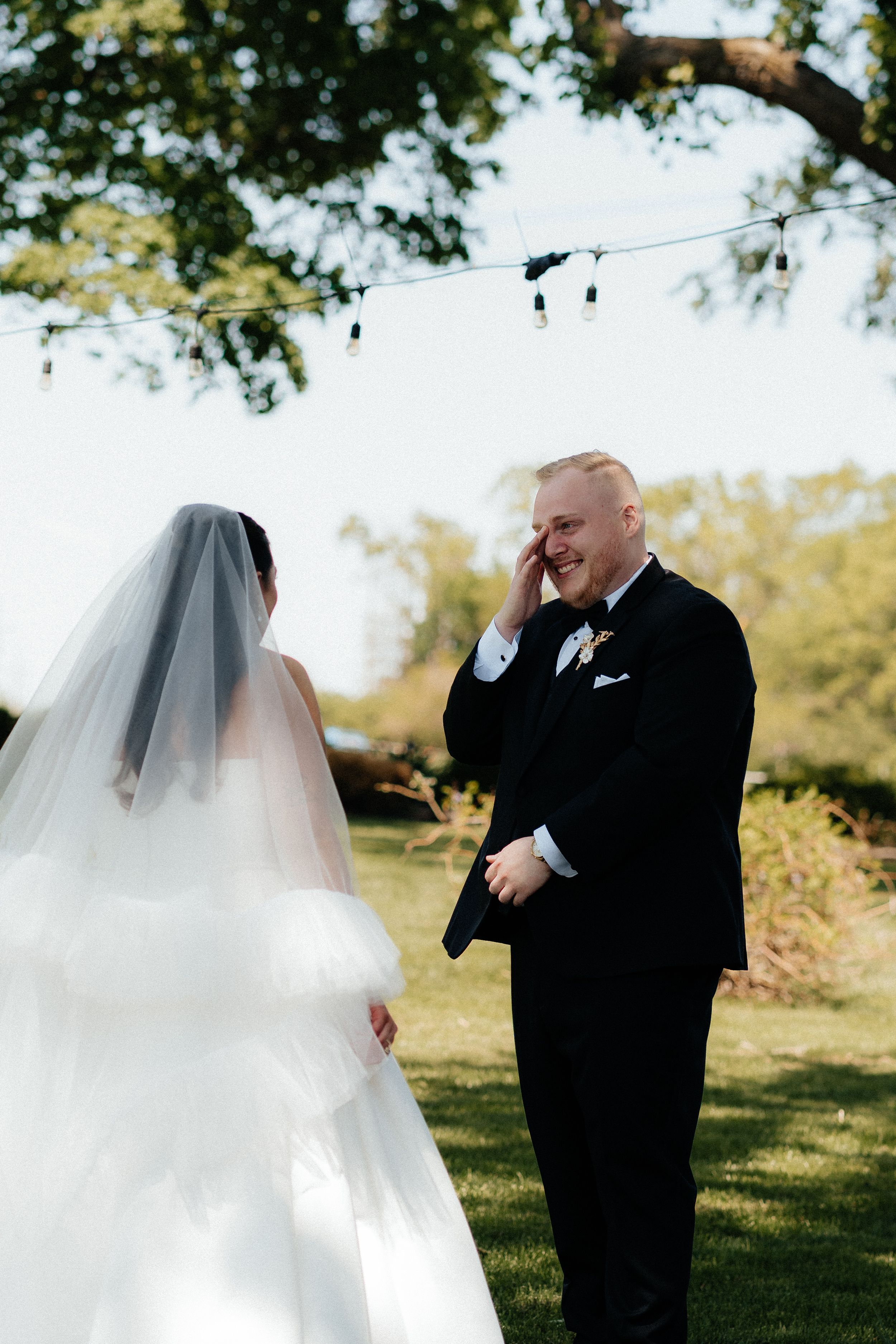 Emotional groom in black tuxedo wipes tear as bride approaches during outdoor first look wedding moment.