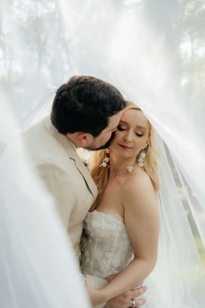 Intimate moment between newlyweds under flowing white veil in soft natural light.