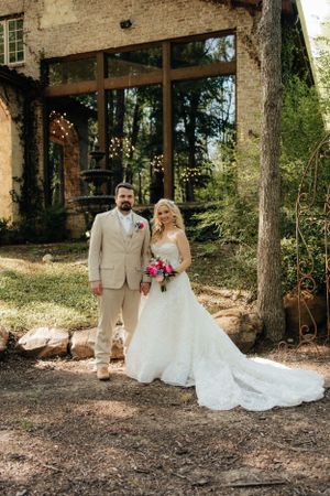 A wedding couple poses in formal attire outside a rustic stone building with trailing greenery.