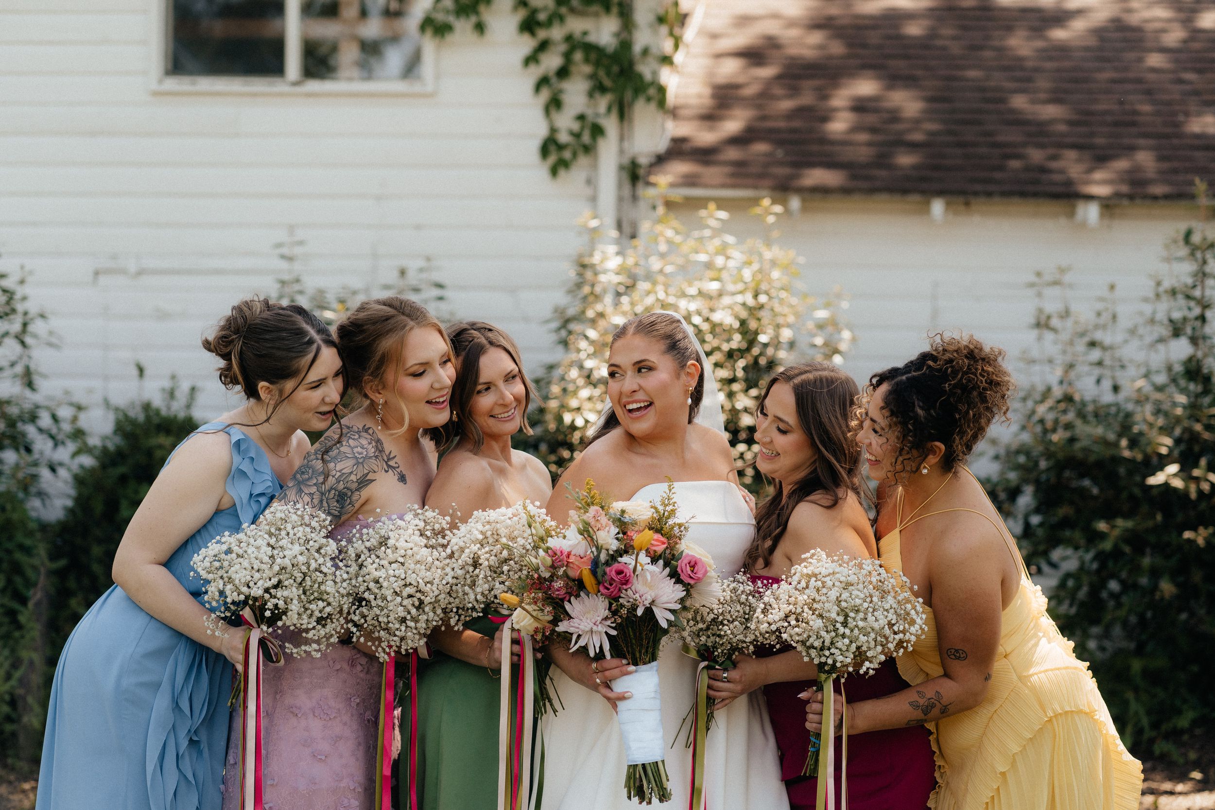 Bride in white gown laughing with colorful bridesmaids holding bouquets outdoors near a white building.
