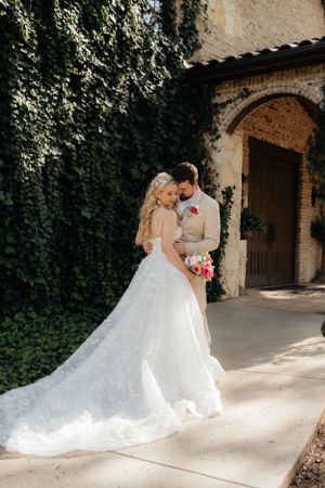 A romantic wedding couple embraces in front of an ivy-covered stone wall and arched doorway.