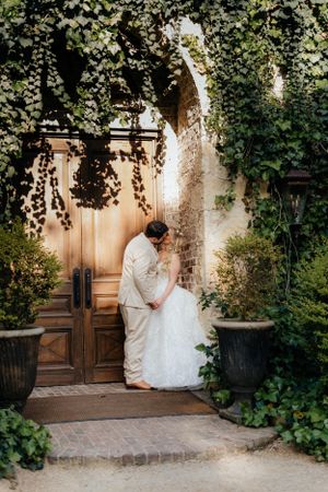 A couple shares an intimate moment by an ornate wooden door surrounded by climbing vines.