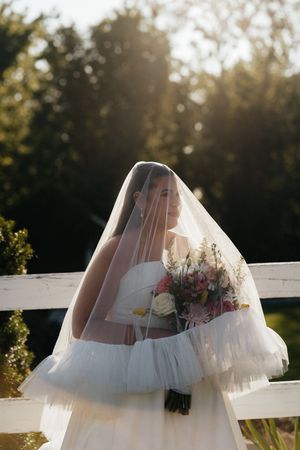 Bride in white tulle gown and veil holds colorful bouquet by white fence in golden sunlight.