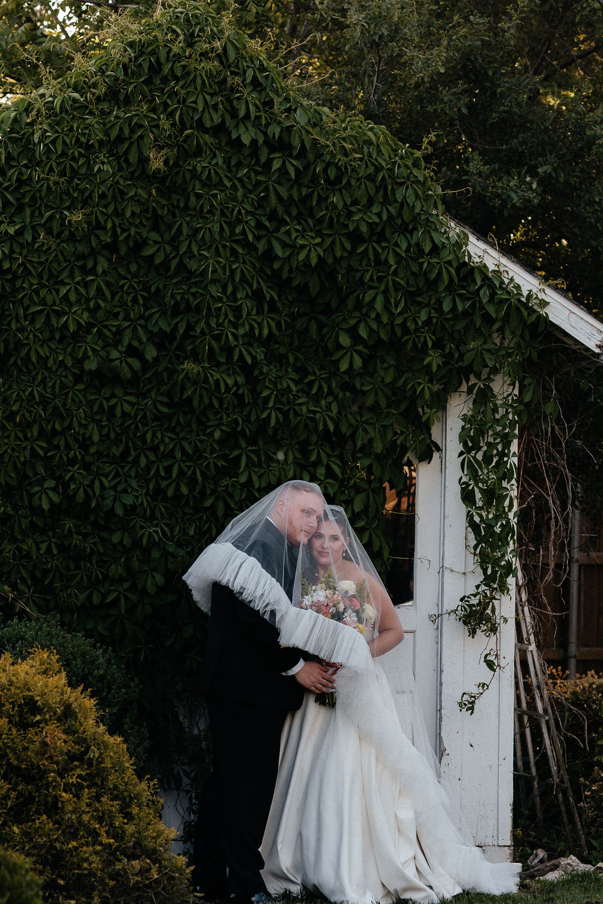 Bride and groom share a romantic moment under her veil beside an ivy-covered white garden shed.