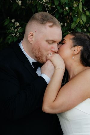 Groom in black tuxedo kisses bride's hand tenderly as she leans in close outdoors.