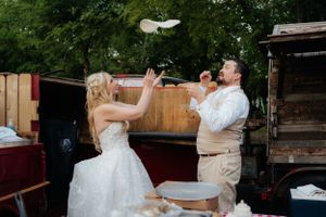 A couple in wedding attire playfully tosses pizza dough in the air during their reception.