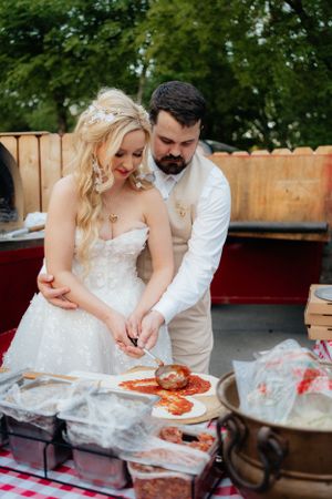 A couple prepares pizza together at their casual outdoor wedding reception.