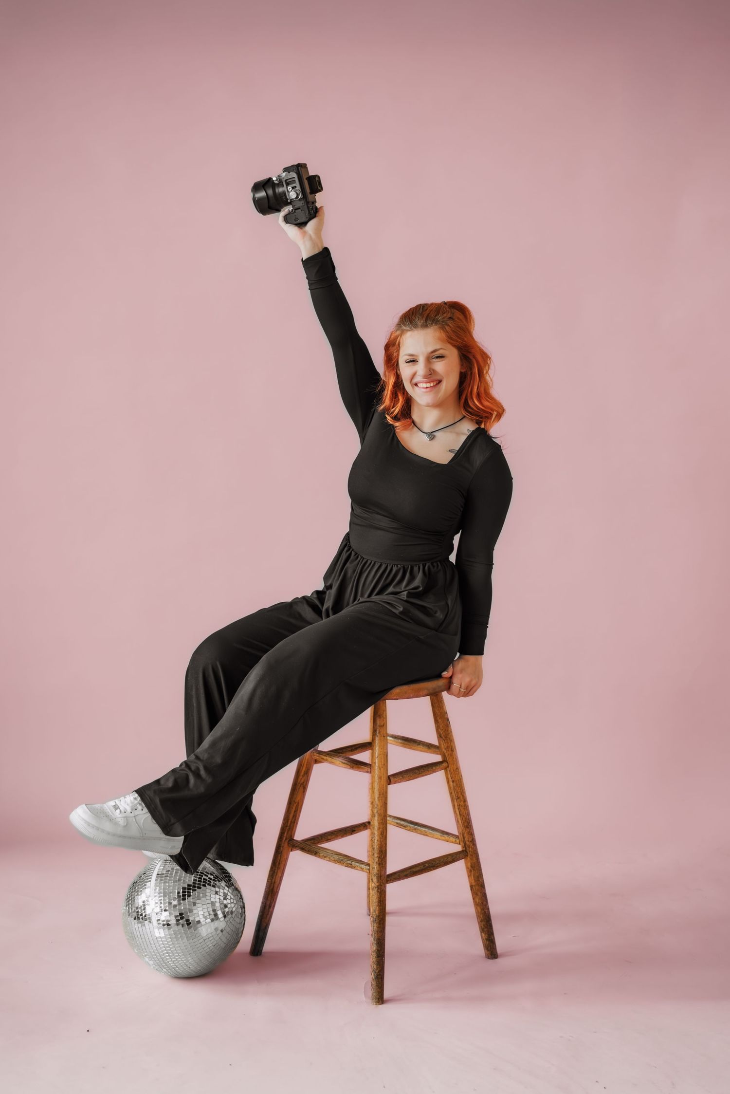 A photographer sits on wooden stool with raised camera and silver disco ball at feet.