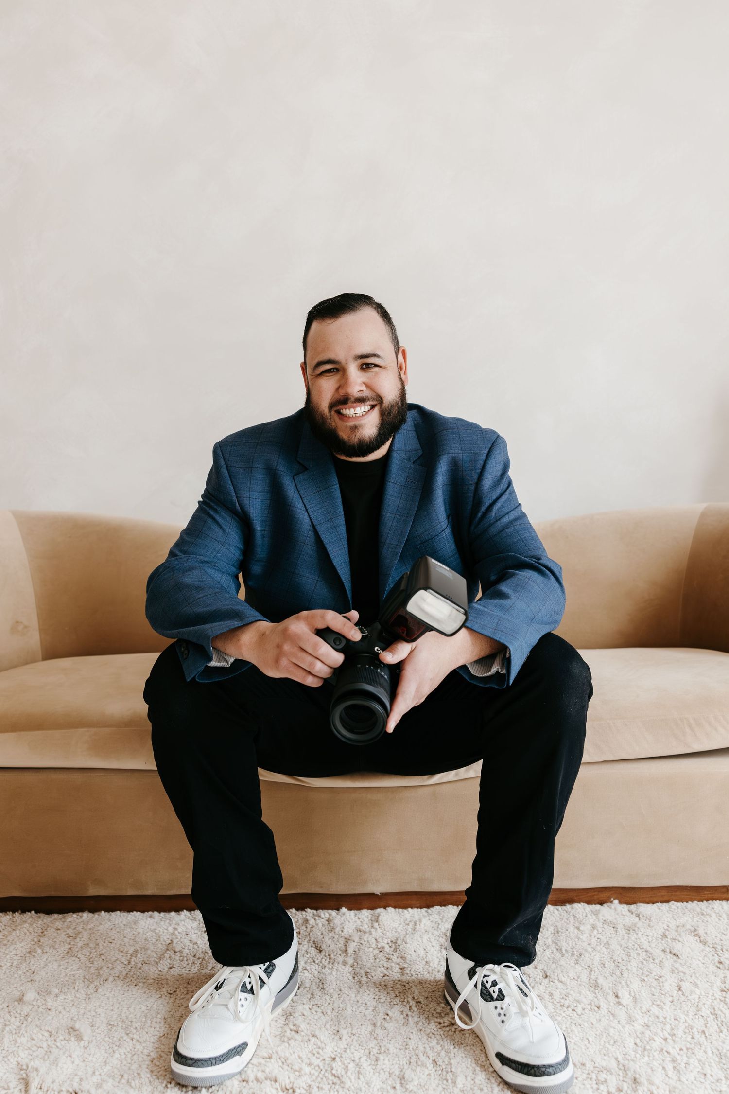Smiling male photographer in blue blazer sits on beige sofa holding professional camera.