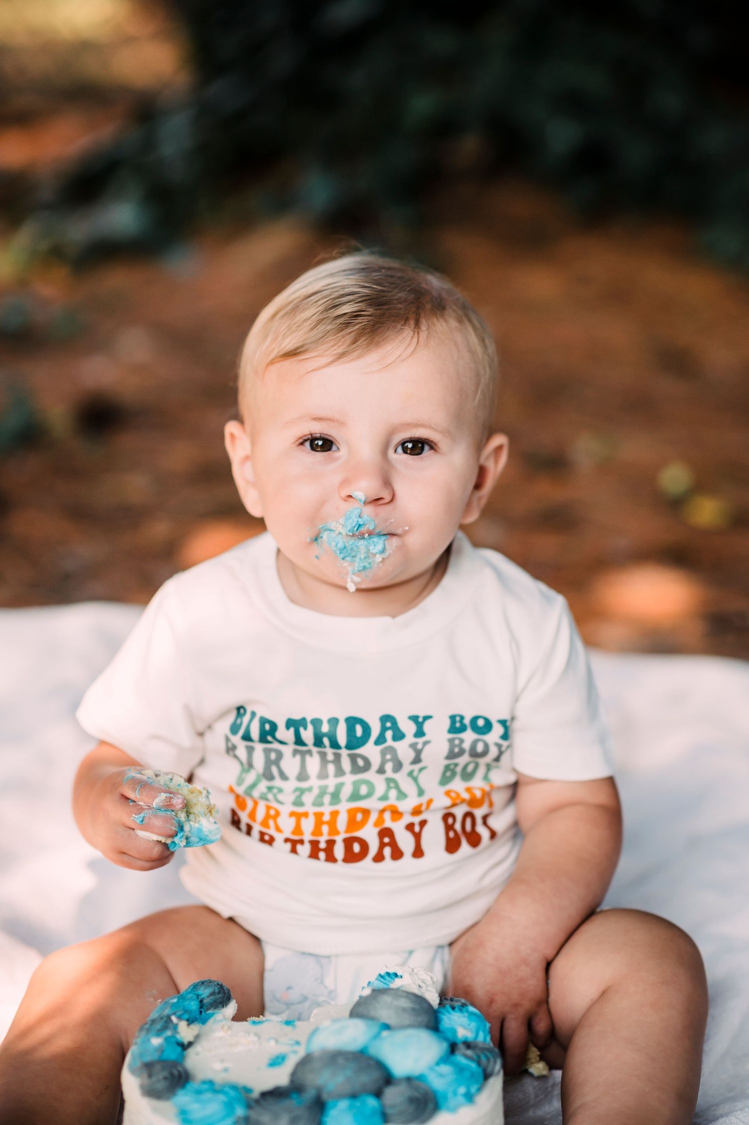 A toddler wearing a birthday boy shirt sits with a blue smeared cake during their first birthday cake smash photo session.