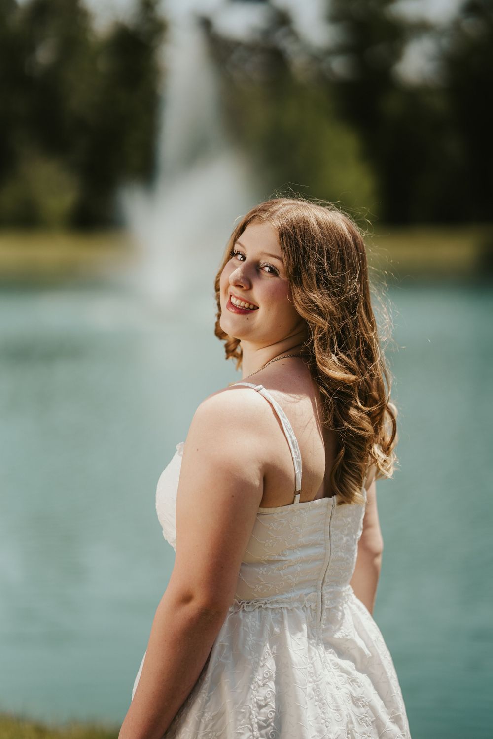 Smiling young woman in white summer dress poses by a fountain in a sunny outdoor setting.