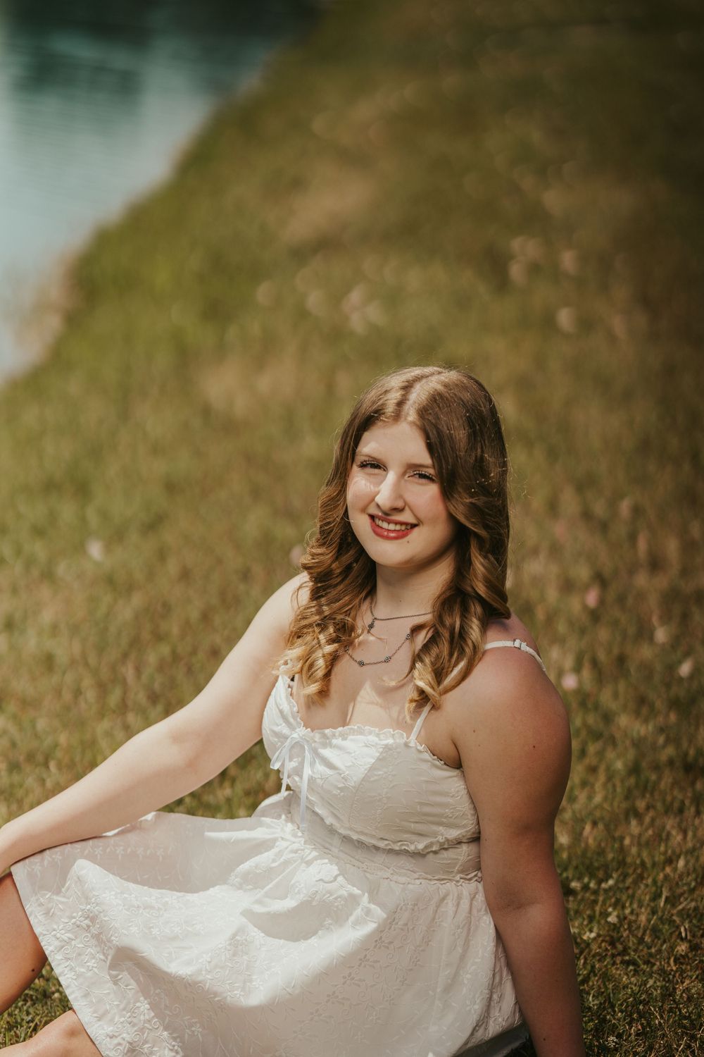 A person in a white sundress sits on grassy hillside during golden hour with a soft dreamy atmosphere.